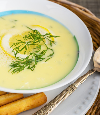 Greek soup avgolemono with a circle of lemon on a white plate on a wicker tray and wooden background with German silver spoon and bread sticks